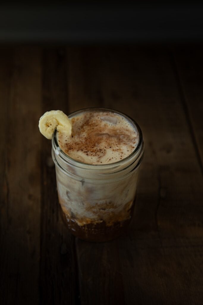 The Korean Vegan: HomeMade 18 A glass jar filled with creamy, homemade iced coffee topped with froth and a sprinkle of cocoa powder. A banana slice graces the rim. This inviting drink rests on a dark, rustic wooden surface, perfect for fans of vegan recipes.