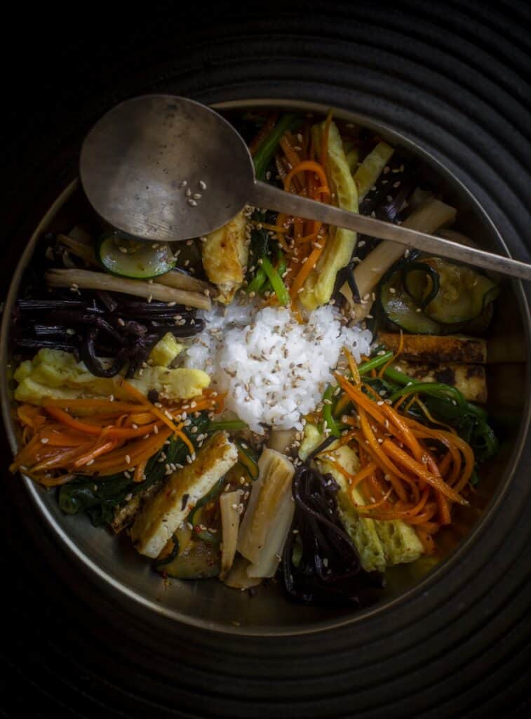 The Korean Vegan: HomeMade 15 A bowl of homemade bibimbap showcases a variety of colorful vegetables, rice, and egg arranged neatly. A metal ladle rests on the rim, with sesame seeds sprinkled on top. The dark background highlights this vibrant Korean vegan dish.