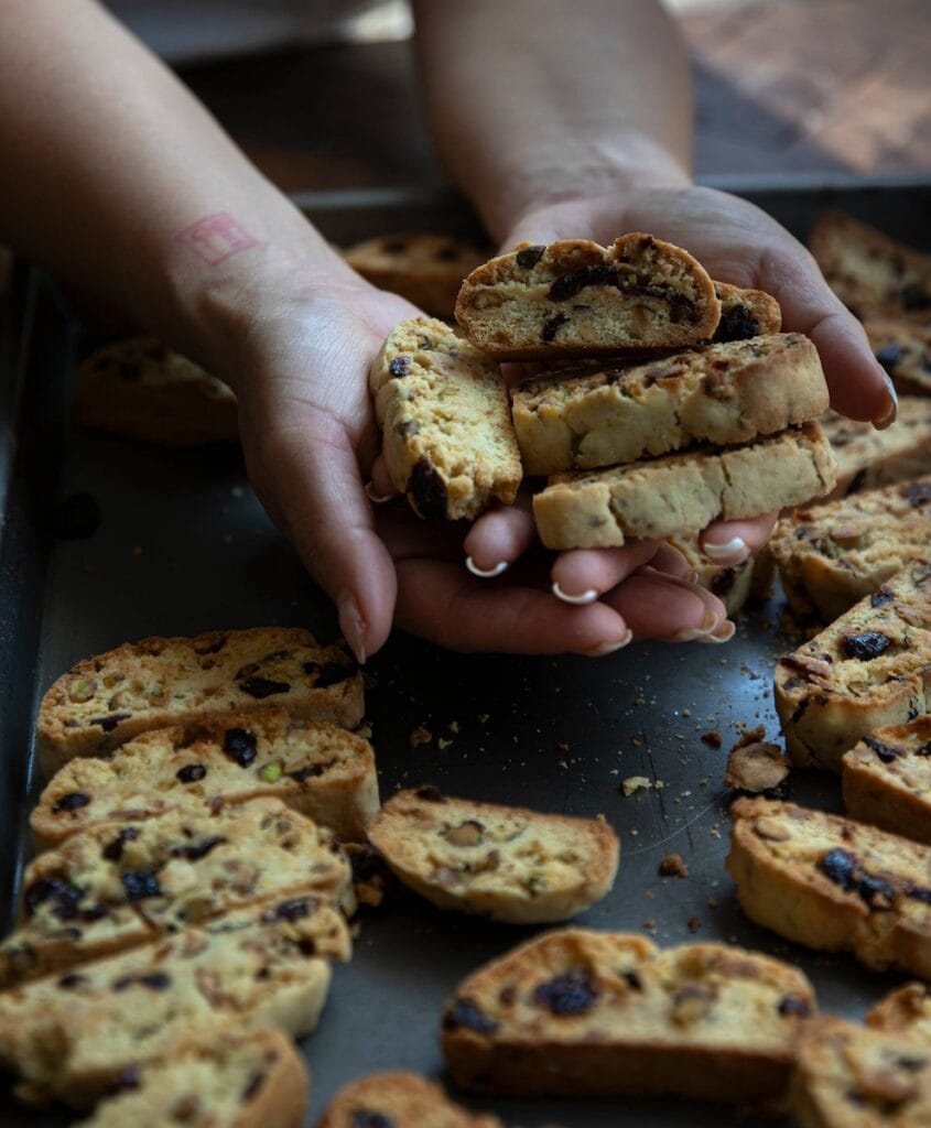 The Korean Vegan: HomeMade 19 Hands hold a homemade batch of rustic biscotti with visible raisins and nuts, showcasing their golden, crunchy texture. Below, more vegan biscotti are scattered on a metal tray, inviting everyone to savor this delectable treat.