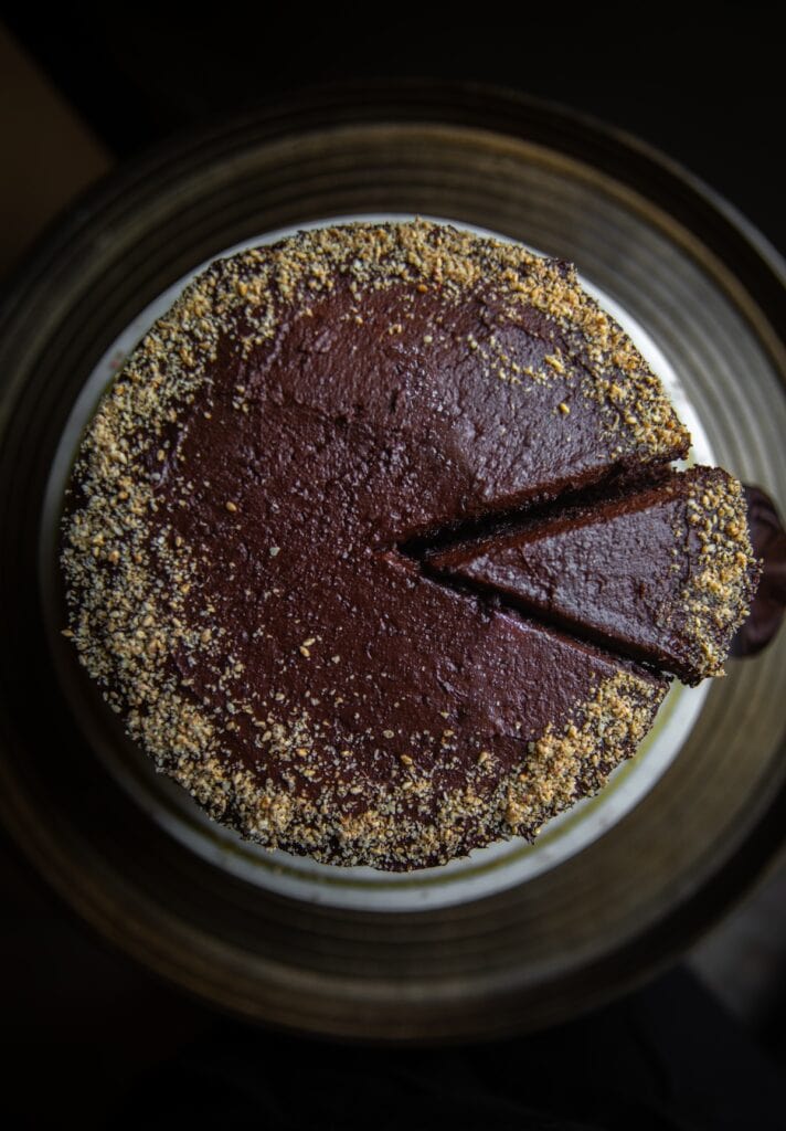 The Korean Vegan: HomeMade 14 A top view of a round, homemade chocolate cake with two slices cut out. The cake is covered in dark frosting and sprinkled with crushed nuts around the edges, sitting on a brown ceramic plate—a delicious treat in the realm of Korean vegan desserts.