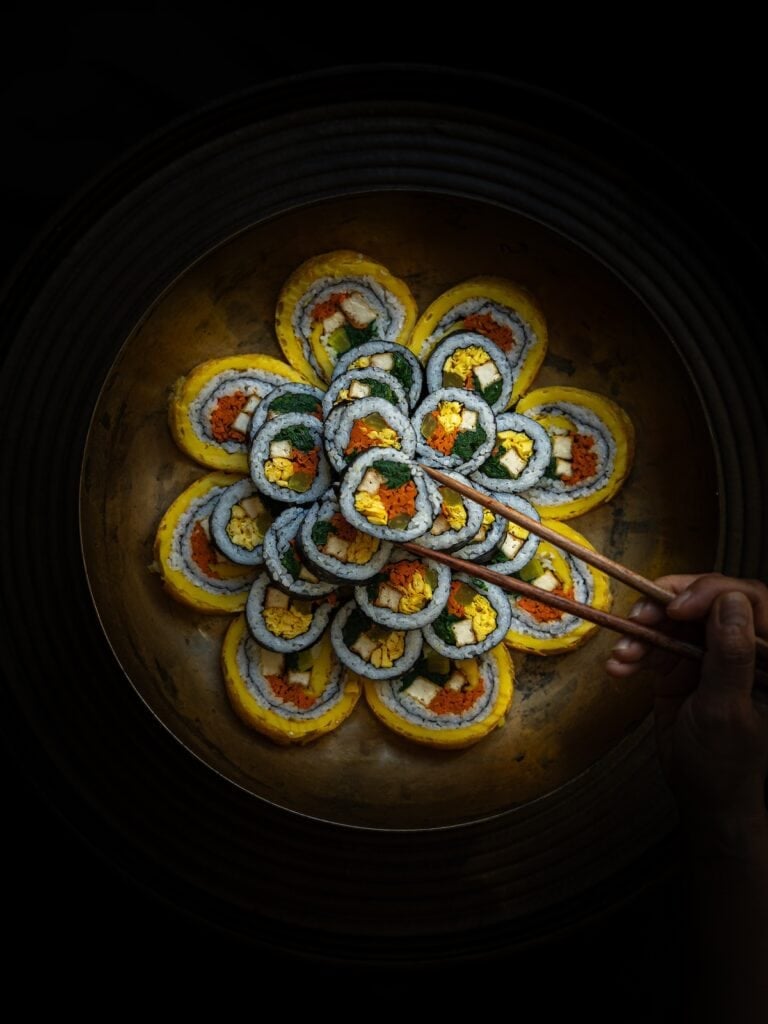 The Korean Vegan: HomeMade 11 A beautifully arranged platter of sushi rolls, crafted with vegan recipes, is displayed in a circular pattern resembling a flower. A hand holds chopsticks, picking up one roll. The lighting highlights the vibrant colors of the ingredients against a dark background.