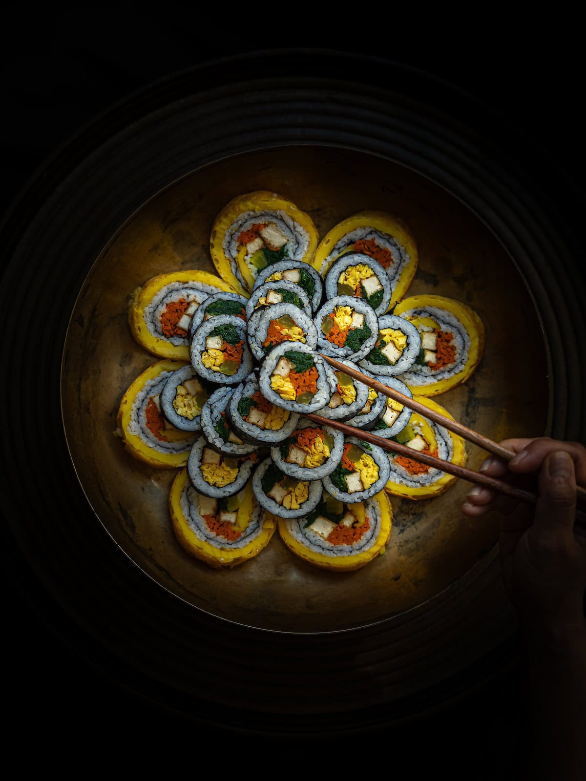 The Korean Vegan: HomeMade 3 A person uses chopsticks to pick up a piece from a circular arrangement of homemade kimbap rolls. These Korean Vegan delights are filled with plant-based egg, carrot, and greens, and displayed on a dark plate. Dim lighting highlights their vibrant colors.