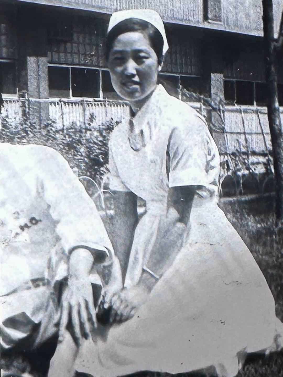 The Korean Vegan: HomeMade 5 A historical black and white photo captures a nurse in a homemade uniform and cap, kneeling outdoors. She smiles warmly in front of a building, with a fenced garden visible in the background.