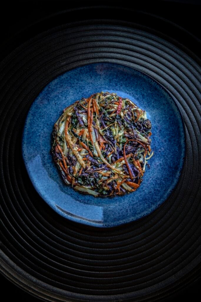 The Korean Vegan: HomeMade 22 A round, homemade vegetable patty with crispy edges rests on a blue textured ceramic plate. Thinly sliced carrots, greens, and other veggies compose this delight, all presented in dark, moody lighting. The plate sits elegantly on a ridged, dark surface.