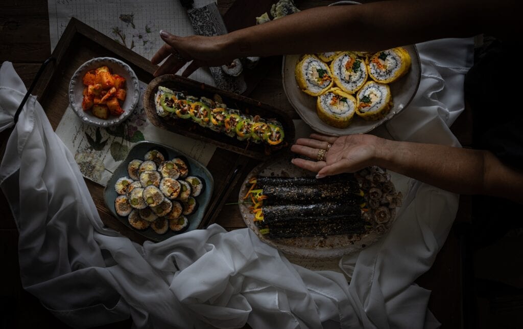 The Korean Vegan: HomeMade 21 A top view of a selection of homemade kimbap on a dark table. A person is thoughtfully arranging the plates, surrounded by white fabric.
