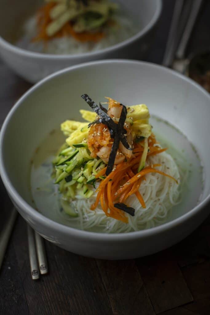 The Korean Vegan: HomeMade 12 A bowl of homemade rice noodles topped with sliced carrots, zucchini, scrambled eggs, nori strips, and a dollop of sauce sits invitingly. Chopsticks rest beside it on the wooden surface. Another similar dish is partially visible in the background, showcasing Korean vegan delights.