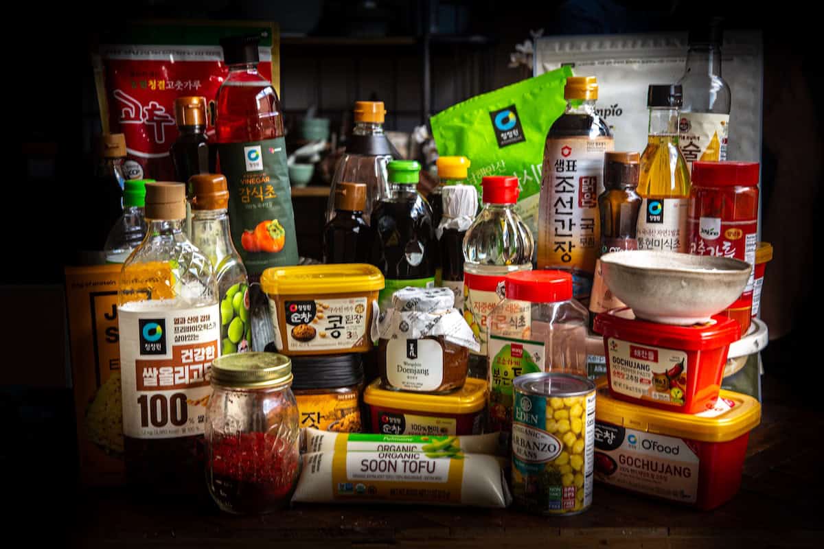 The Korean Vegan: HomeMade 4 A collection of various Korean vegan food products including sauces, oils, tofu, and rice. Containers of different sizes and colors are arranged closely together on a wooden surface.