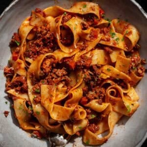 BEST Vegan Bolognese Recipe - Meaty and Delicious. 41 A bowl of wide, ribbon-like pasta topped with chunky vegan bolognese sauce, garnished with chopped herbs, and served on a gray plate.