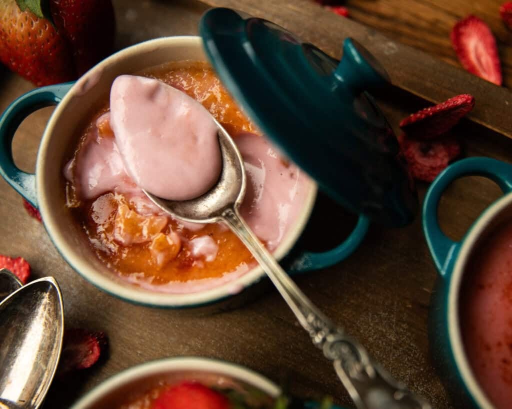 Pink Strawberry Crème Brûlée (Vegan & Easy) 8 A small teal ceramic dish with a lid holds creamy pink Strawberry Crème Brûlée. A spoon scoops up some of the dessert, while fresh strawberries and another dish rest nearby on the wooden surface.