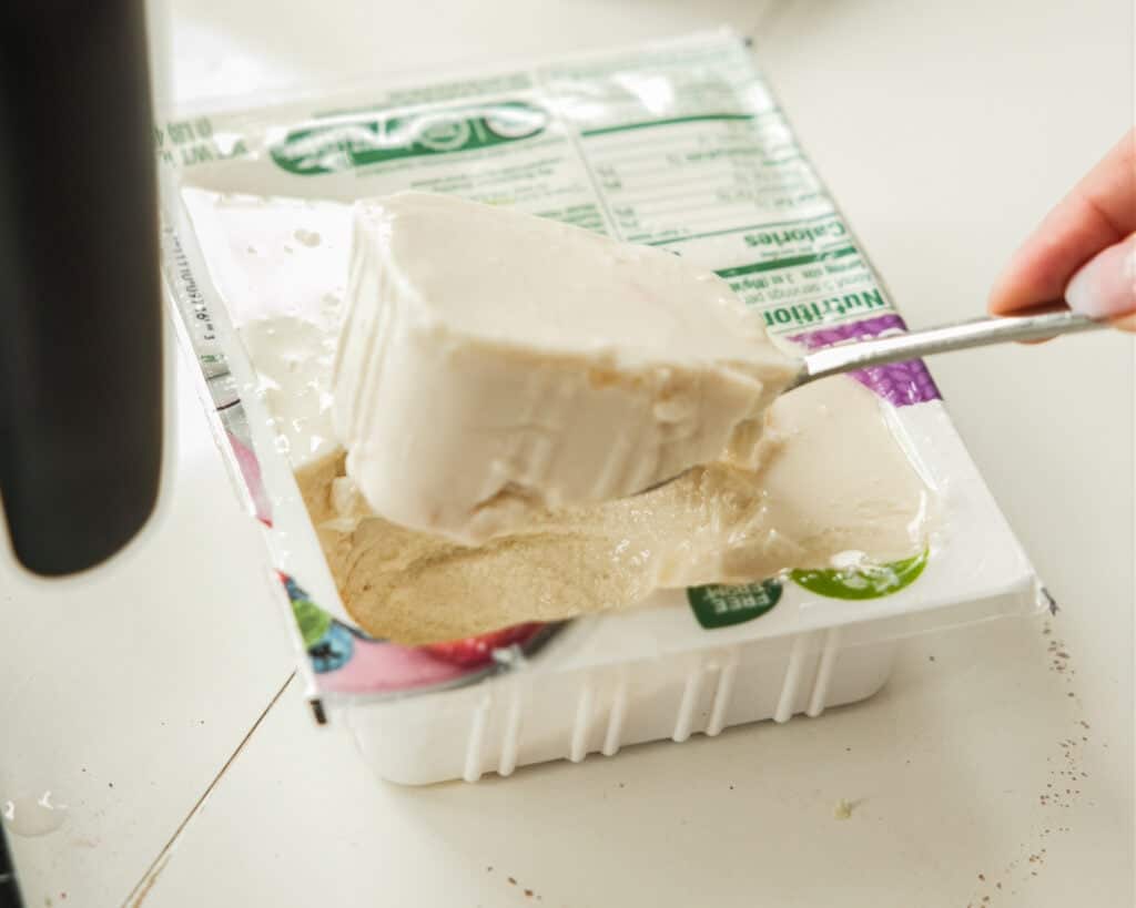 Pink Strawberry Crème Brûlée (Vegan & Easy) 2 A hand scoops a large piece of soft tofu from an open plastic container with a spoon. The tofu looks smooth and creamy—perfect for making Vegan Crème Brûlée—and the nutritional label is partially visible on the packaging.