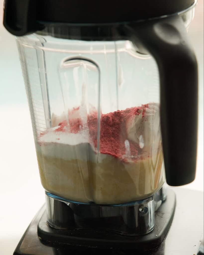 Pink Strawberry Crème Brûlée (Vegan & Easy) 9 A close-up of a blender filled with yellow liquid, white powder, and a layer of red powder, ready to be mixed for an easy Strawberry Crème Brûlée.