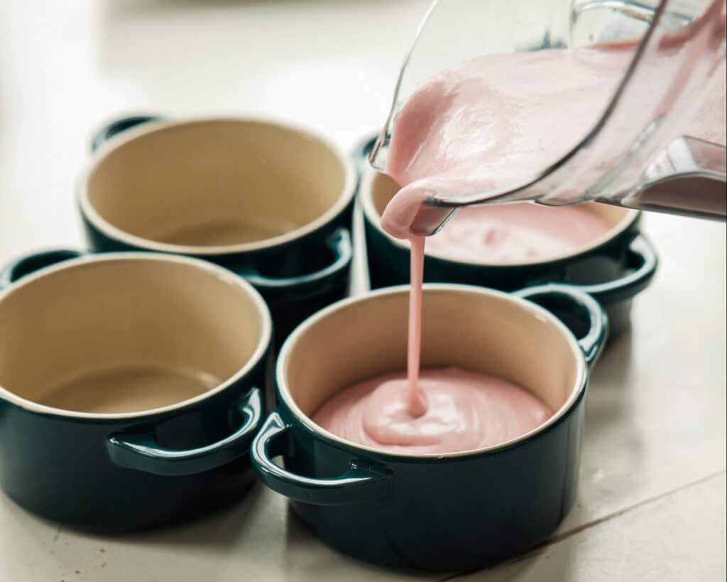 Pink Strawberry Crème Brûlée (Vegan & Easy) 11 A pink vegan Strawberry Crème Brûlée mixture is being poured from a blender into a small round ceramic dish. Three similar empty dishes sit beside it on a white surface, showcasing an easy dessert prep.