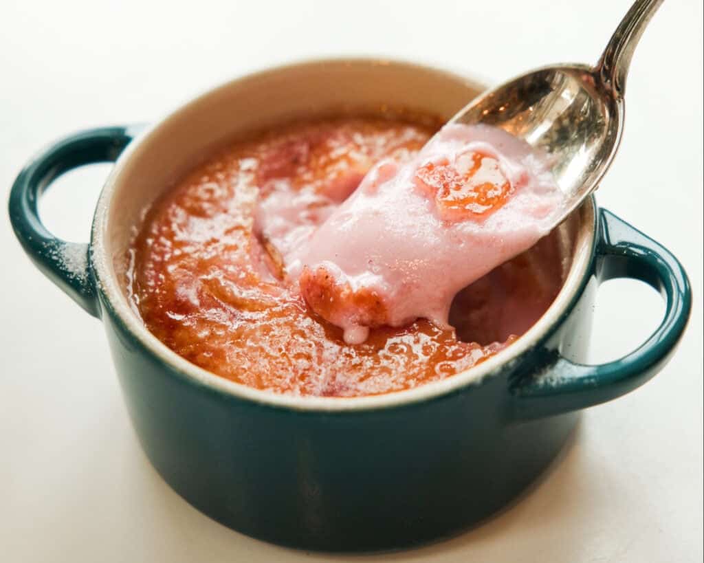 Pink Strawberry Crème Brûlée (Vegan & Easy) 15 A spoon scoops up pink, creamy Strawberry Crème Brûlée from a small, round, blue baking dish with handles, revealing a smooth golden-brown top layer and a soft, airy interior—an easy recipe for an elegant dessert.