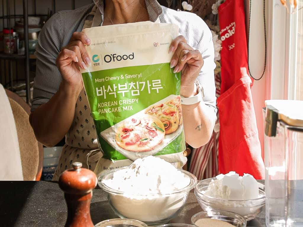 Easy and Crispy Korean Kimchi Pancakes (김치전) Recipe. 5 A person in an apron holds a bag of OFood Korean Crispy Pancake Mix over a kitchen counter with bowls of flour, a pepper grinder, and kitchen tools in sunlight.