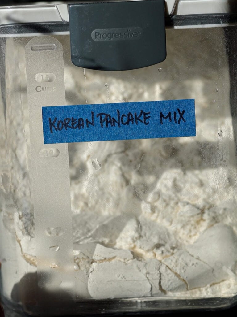 Easy and Crispy Korean Kimchi Pancakes (김치전) Recipe. 4 A close-up of a plastic container labeled Korean Pancake Mix with blue tape. The container is filled with white powder and has a measurement strip on the side.
