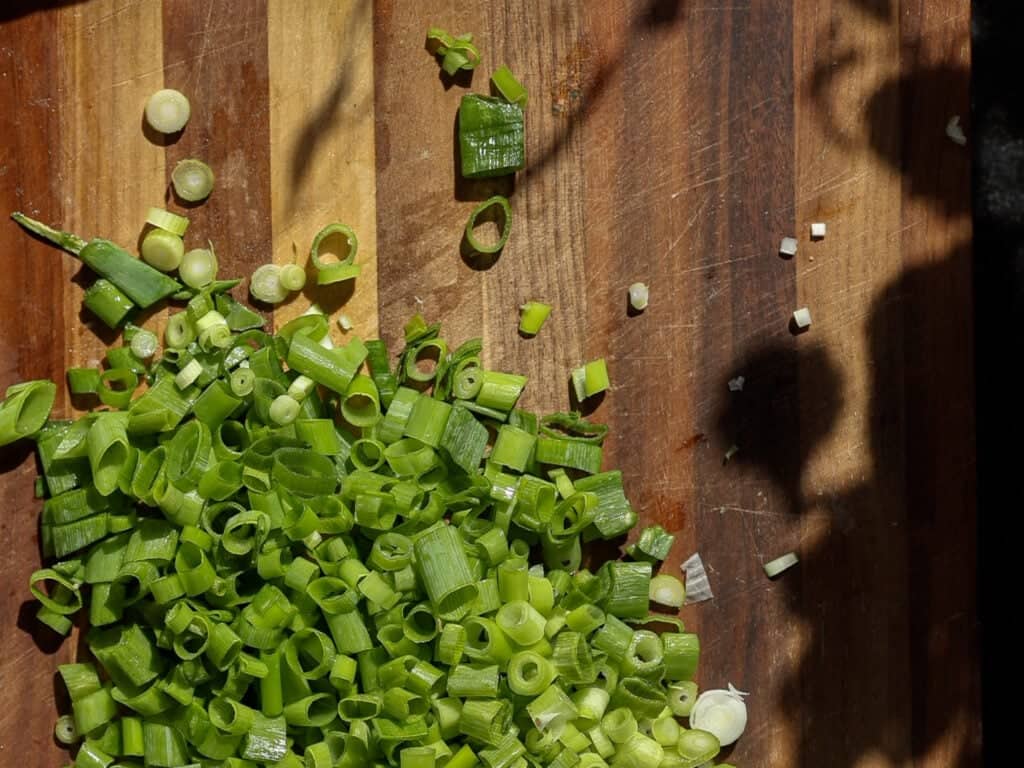 Easy and Crispy Korean Kimchi Pancakes (김치전) Recipe. 9 Chopped green onions scattered on a wooden cutting board, with some shadows cast across the surface.