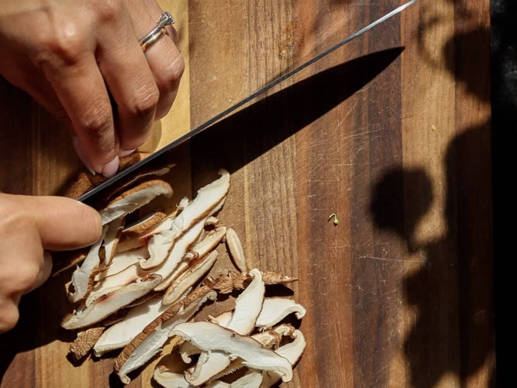Easy and Crispy Korean Kimchi Pancakes (김치전) Recipe. 8 A person’s hands slice shiitake mushrooms on a wooden cutting board with a large knife, creating thin, even pieces. Sunlight and shadows fall across the scene.