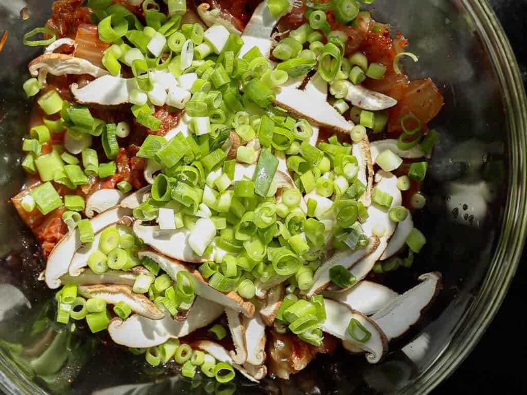 Easy and Crispy Korean Kimchi Pancakes (김치전) Recipe. 12 A glass bowl filled with chopped green onions, sliced mushrooms, and kimchi, viewed from above. Bright sunlight highlights the fresh ingredients, emphasizing their colors and textures.