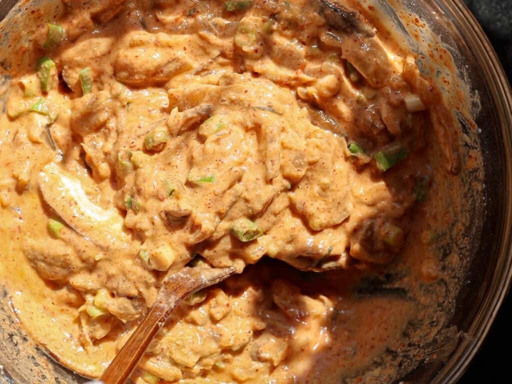 Easy and Crispy Korean Kimchi Pancakes (김치전) Recipe. 14 A close-up of a bowl filled with a thick, orange-tinted mixture containing chopped green herbs or vegetables, with a wooden spoon partially submerged in the mixture.