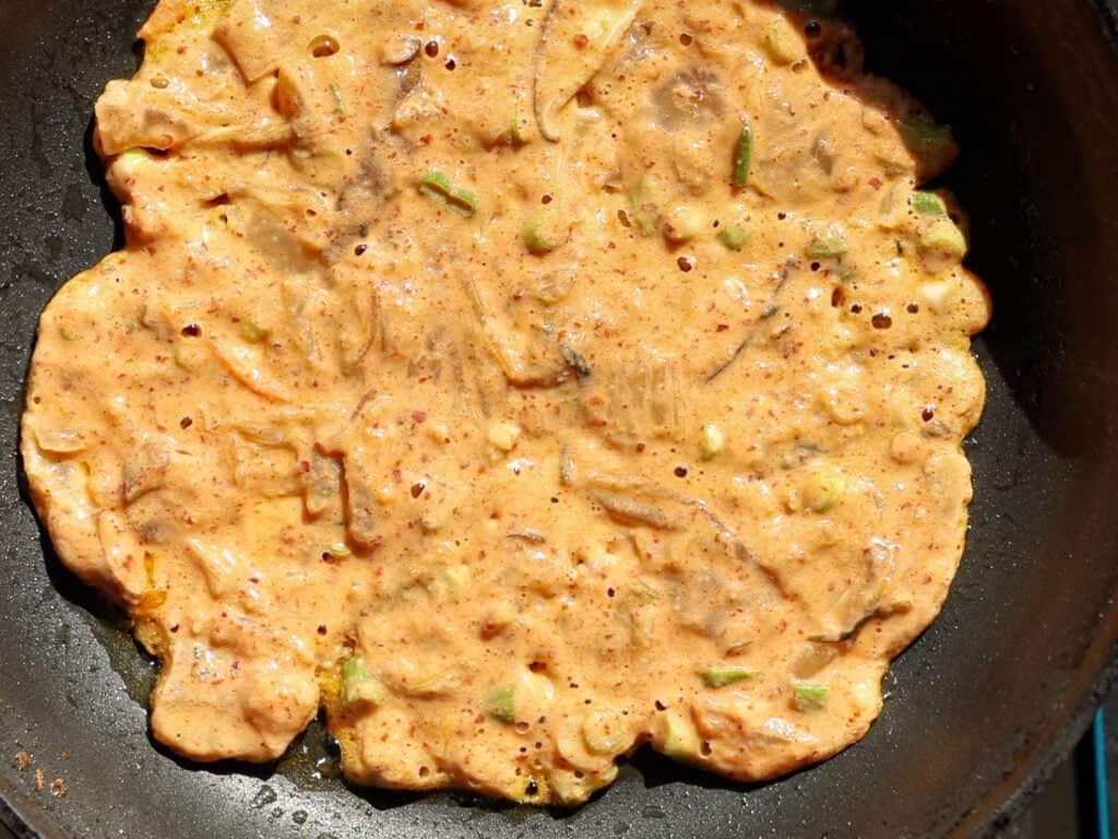 Easy and Crispy Korean Kimchi Pancakes (김치전) Recipe. 16 A close-up of a Korean pancake (jeon) cooking in a black pan, with visible bits of vegetables in the orange batter. The pancake is partially cooked, with some bubbles and uneven edges.