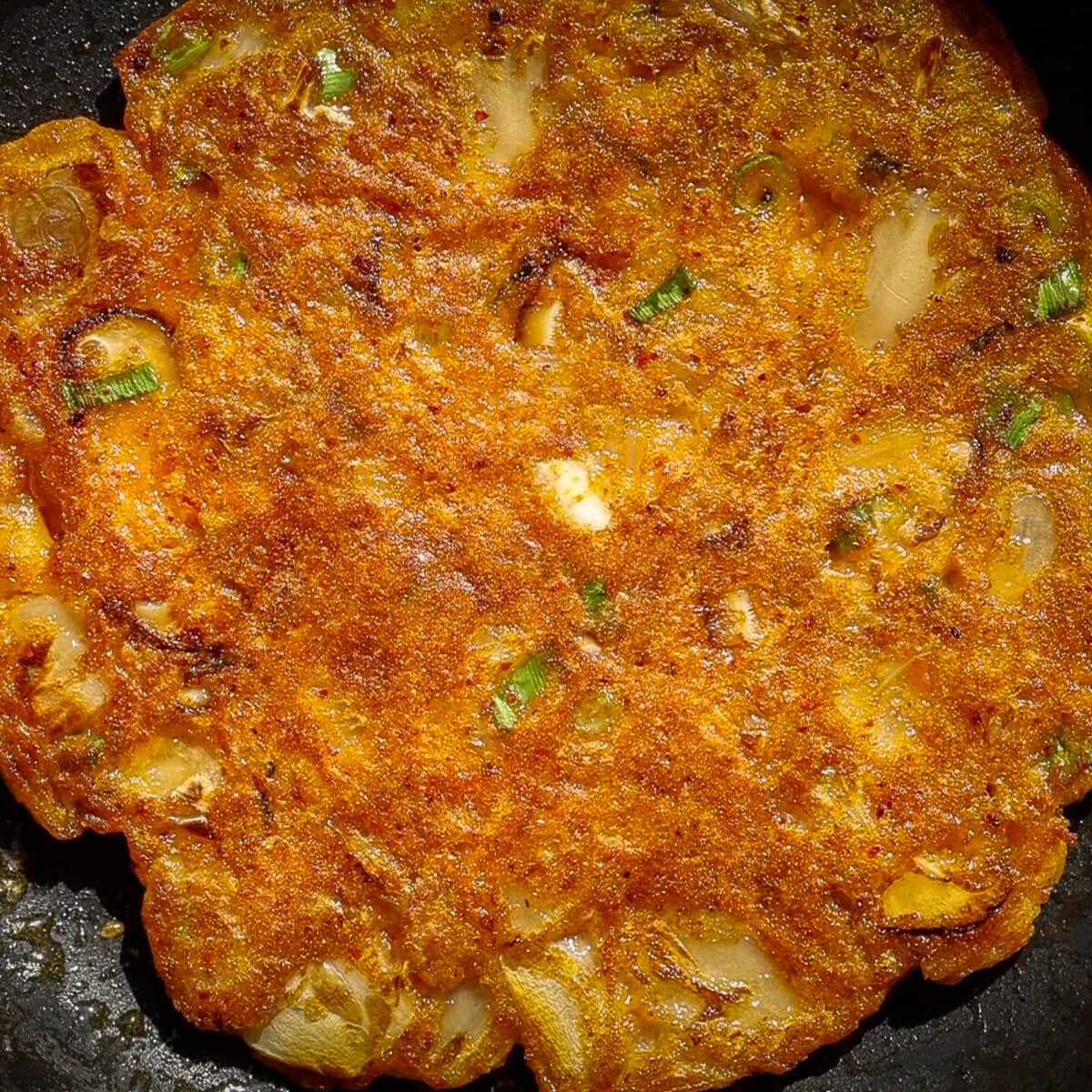 10+ Delicious and Easy Recipes Using Kimchi Today! 7 A close-up of golden-brown, crispy Korean kimchi pancakes with visible pieces of green onion and other vegetables cooked into the surface.
