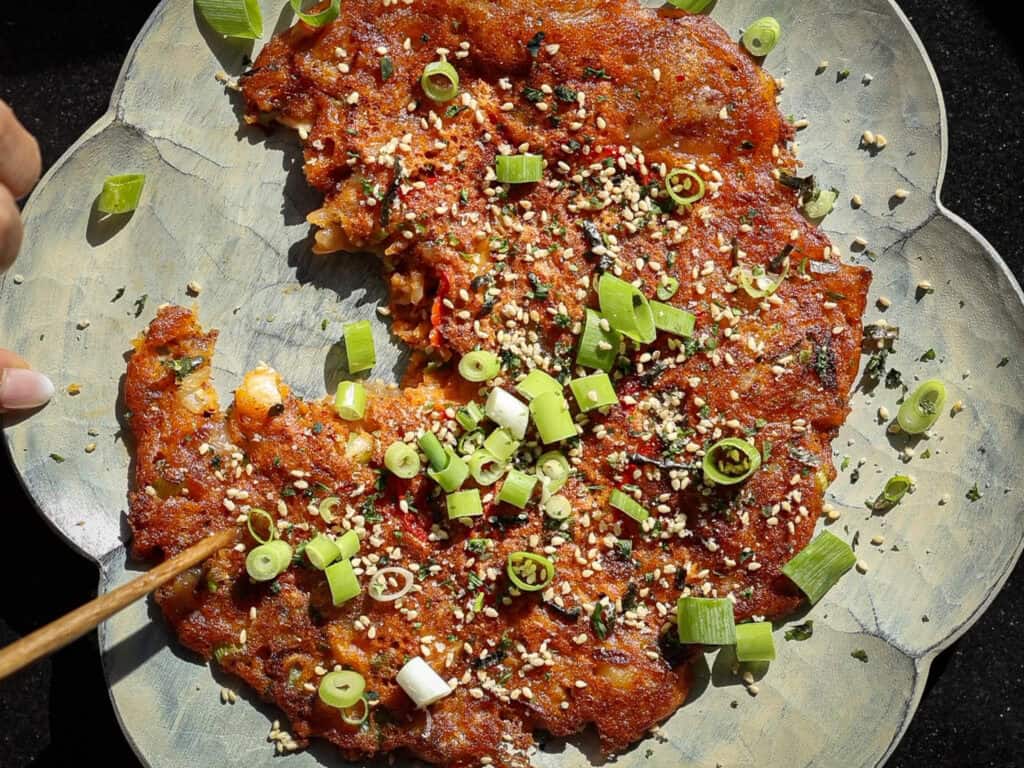 Easy and Crispy Korean Kimchi Pancakes (김치전) Recipe. 2 A savory kimchi pancake garnished with chopped green onions, sesame seeds, and herbs is served on a decorative plate. A hand is using chopsticks to take a piece from the crispy, golden-brown pancake.