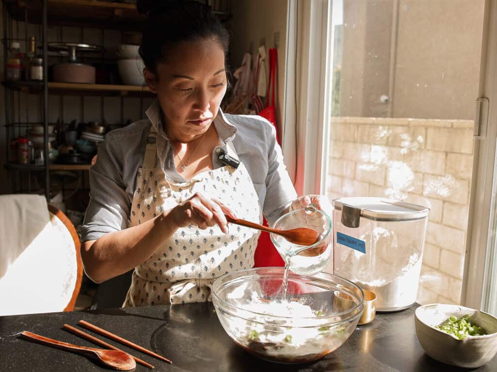 Easy and Crispy Korean Kimchi Pancakes (김치전) Recipe. 13 A woman wearing an apron pours water into a mixing bowl of flour while preparing food in a sunlit kitchen, with cooking utensils and ingredients on the table.