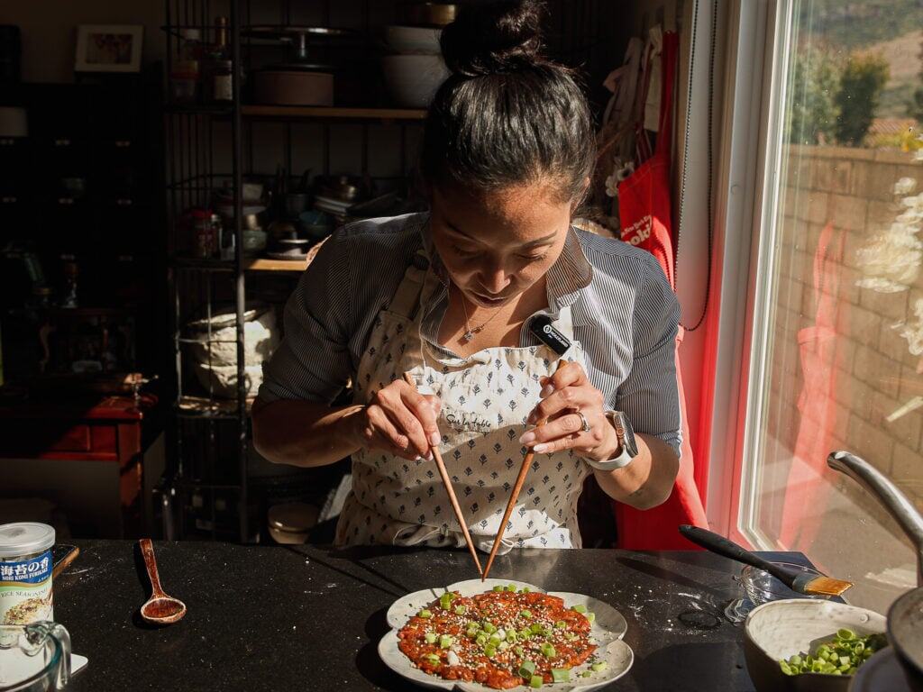 Easy and Crispy Korean Kimchi Pancakes (김치전) Recipe. 18 A woman wearing an apron uses chopsticks to garnish a dish with green onions in a sunlit kitchen, standing by a counter near a window.