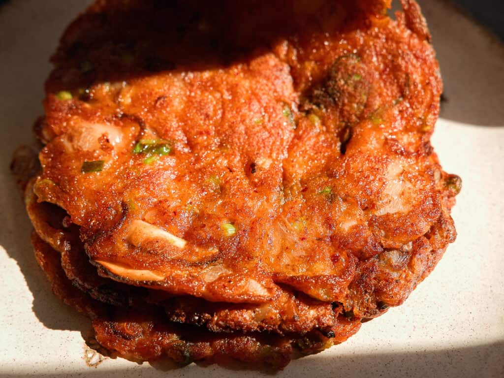 Easy and Crispy Korean Kimchi Pancakes (김치전) Recipe. 3 A close-up of crispy, golden-brown Korean kimchi pancakes stacked on a plate, with visible bits of cabbage and green onions, lit by natural sunlight.