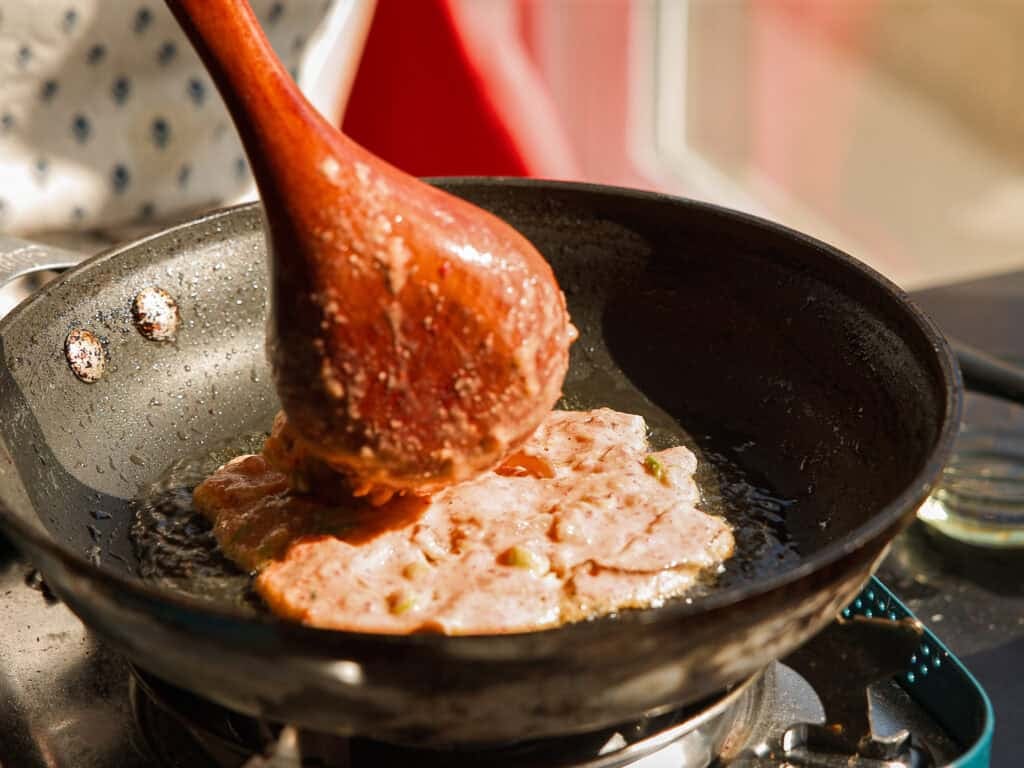 Easy and Crispy Korean Kimchi Pancakes (김치전) Recipe. 15 A person uses a wooden spoon to spread kimchi pancake mixture in a frying pan on a stovetop, with sunlight illuminating the scene.