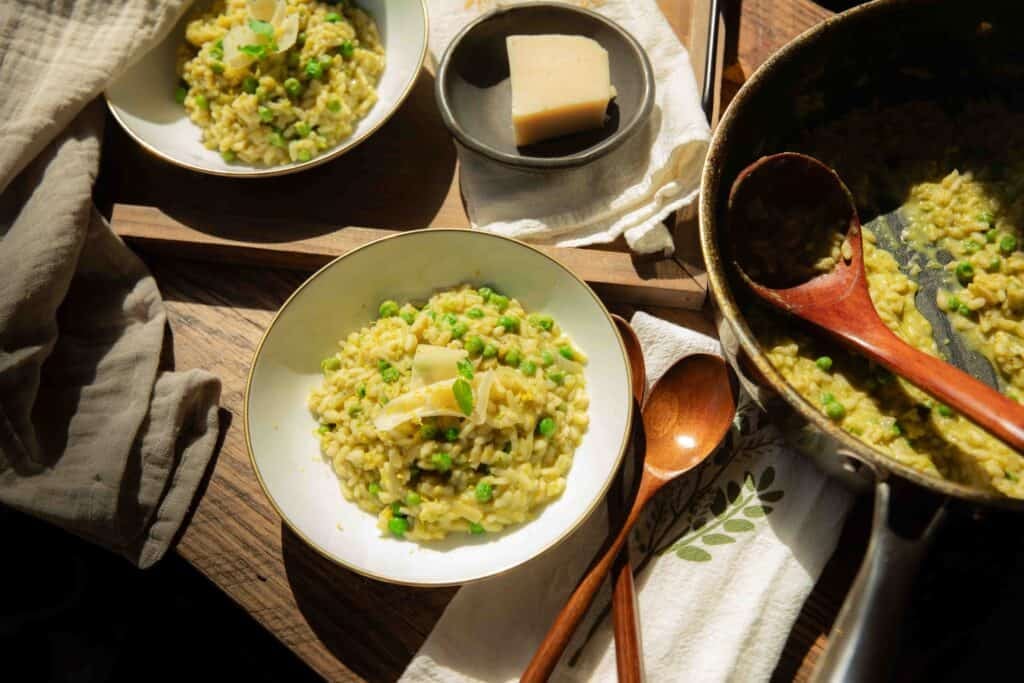 The BEST Sweet Green Pea Risotto Recipe (Vegan). 21 Two bowls of creamy green pea risotto with cheese sit on a wooden table, next to a skillet filled with more risotto, a wooden spoon, a linen napkin, and a small dish holding a block of cheese.