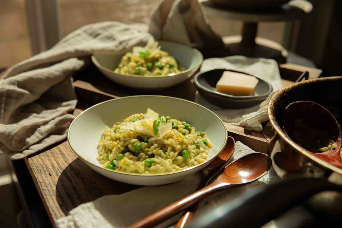 The BEST Sweet Green Pea Risotto Recipe (Vegan). 1 Two bowls of creamy green pea risotto with green peas and cheese sit on a tray, accompanied by a small plate with a wedge of cheese, wooden spoons, and a beige napkin in warm, natural sunlight.