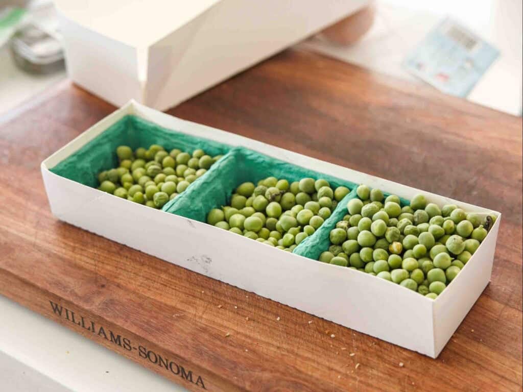 The BEST Sweet Green Pea Risotto Recipe (Vegan). 5 A rectangular white box with three compartments, each filled with green peas, sits on a wooden cutting board labeled WILLIAMS-SONOMA. The box is lined with blue-green paper.