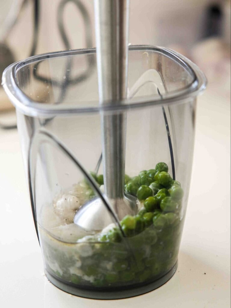 The BEST Sweet Green Pea Risotto Recipe (Vegan). 10 A handheld immersion blender is inside a clear plastic container, blending green peas and chopped onions on a kitchen counter.