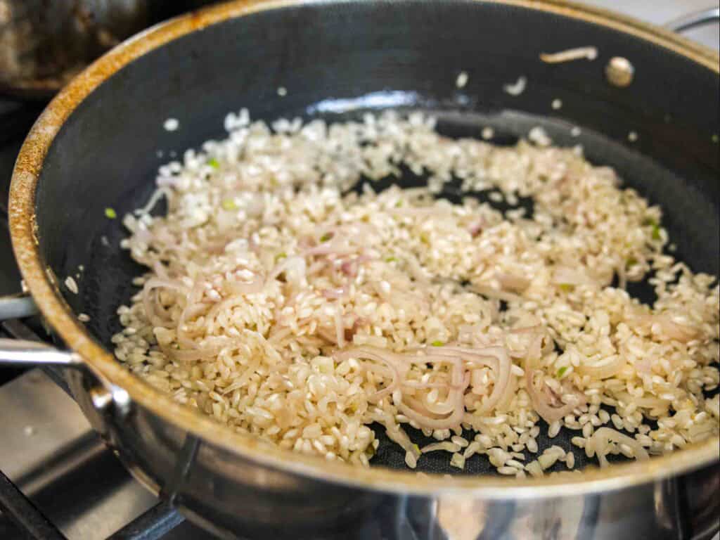 The BEST Sweet Green Pea Risotto Recipe (Vegan). 13 A close-up of a pan on a stove with Arborio rice and sliced shallots sautéing, beginning the process of making risotto. The ingredients are lightly browned and glistening in the pan.