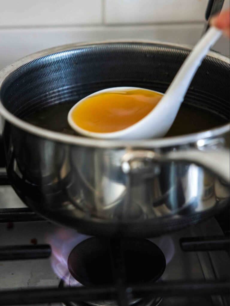 The BEST Sweet Green Pea Risotto Recipe (Vegan). 8 A white ladle scooping clear golden broth from a stainless steel pot on a stove with a visible blue flame beneath.
