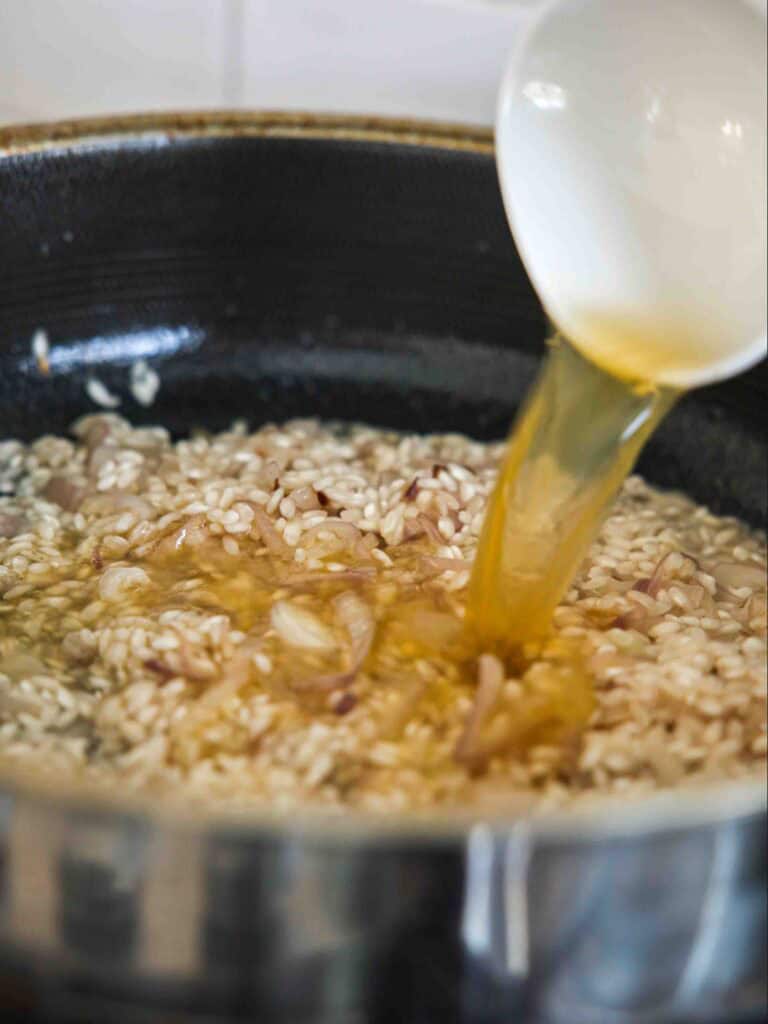 The BEST Sweet Green Pea Risotto Recipe (Vegan). 16 A close-up of a ladle pouring broth into a pan of rice, likely in the process of making risotto, with visible grains and pieces of onion or shallot.