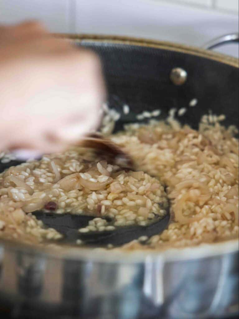 The BEST Sweet Green Pea Risotto Recipe (Vegan). 17 A close-up of a person stirring creamy risotto in a black pan on the stove, with visible grains of rice and caramelized onions.