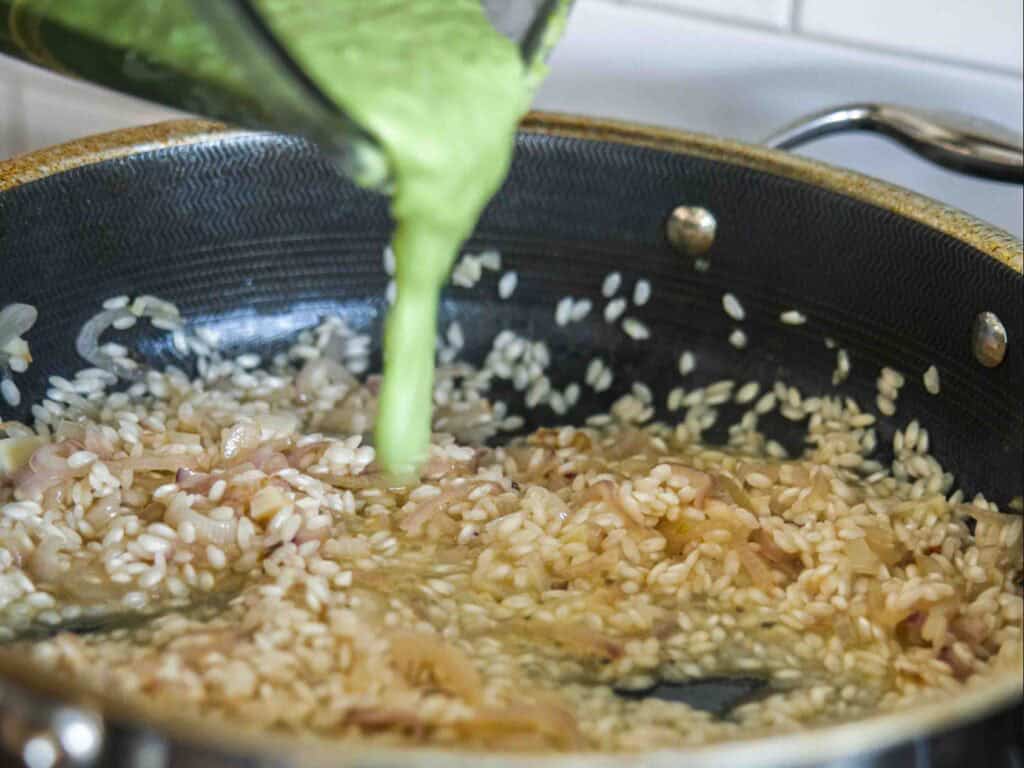The BEST Sweet Green Pea Risotto Recipe (Vegan). 18 A close-up of a pan with sautéed onions and uncooked rice, while a green liquid is being poured in from a blender, likely for a risotto recipe.