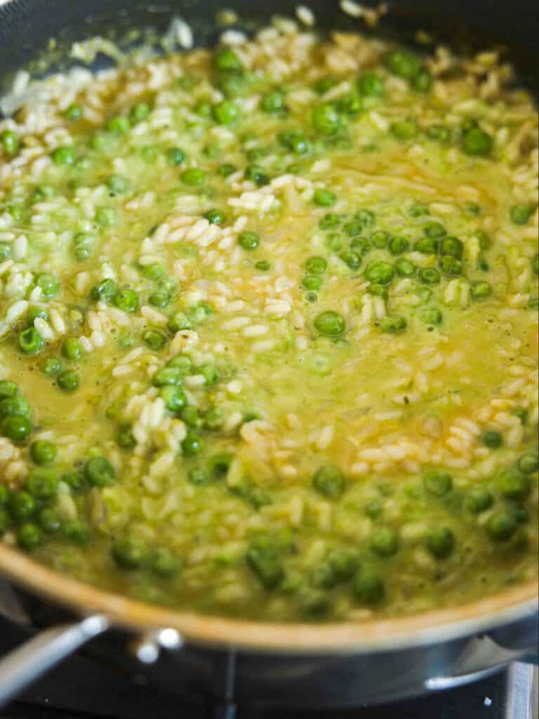 The BEST Sweet Green Pea Risotto Recipe (Vegan). 20 A close-up of a pan filled with creamy risotto, featuring green peas and rice simmering in a yellowish broth.