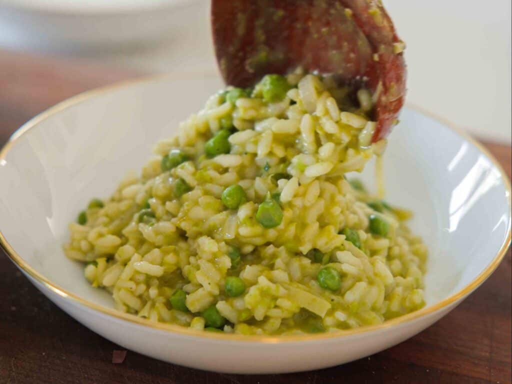 The BEST Sweet Green Pea Risotto Recipe (Vegan). 19 A close-up of creamy risotto with green peas being served onto a white plate with a gold rim using a wooden spoon.