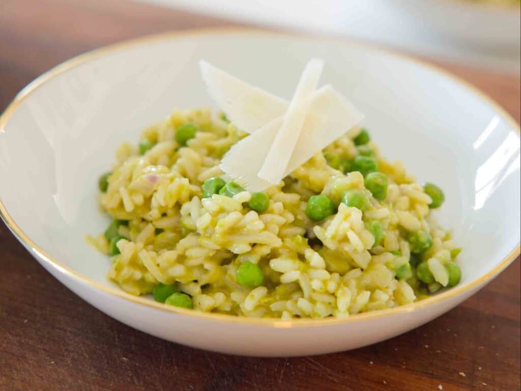 The BEST Sweet Green Pea Risotto Recipe (Vegan). 2 A bowl of creamy green pea risotto with green peas, topped with three thin shavings of Parmesan cheese, served in a white bowl with a gold rim on a wooden surface.