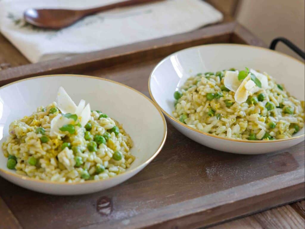 The BEST Sweet Green Pea Risotto Recipe (Vegan). 3 Two bowls of creamy green pea risotto with green peas and topped with cheese shavings, served on a wooden tray. A wooden spoon and a cloth napkin are visible in the background.