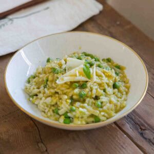 The BEST Sweet Green Pea Risotto Recipe (Vegan). 22 A white bowl with a gold rim holds creamy risotto mixed with green peas and topped with cheese shavings and herbs. The bowl sits on a rustic wooden table next to a white napkin.