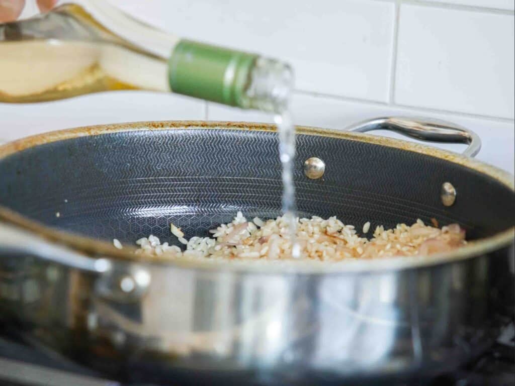 The BEST Sweet Green Pea Risotto Recipe (Vegan). 14 A close-up of a pan on a stove with chopped onions and rice being cooked for green pea risotto, while white wine is being poured in from a bottle.