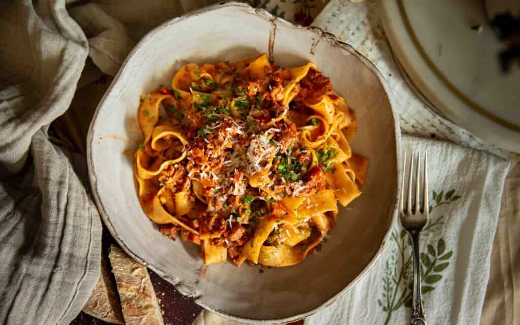 BEST Vegan Bolognese Recipe - Meaty and Delicious. 13 A bowl of wide ribbon pasta topped with vegan bolognese, grated cheese, and herbs sits on a table with a fork, napkins, and slices of bread nearby.