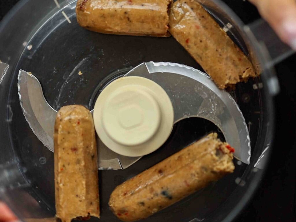BEST Vegan Bolognese Recipe - Meaty and Delicious. 24 Four vegan sausages placed in a food processor, viewed from above, ready to be blended. The sausages have visible herbs and spices within them.