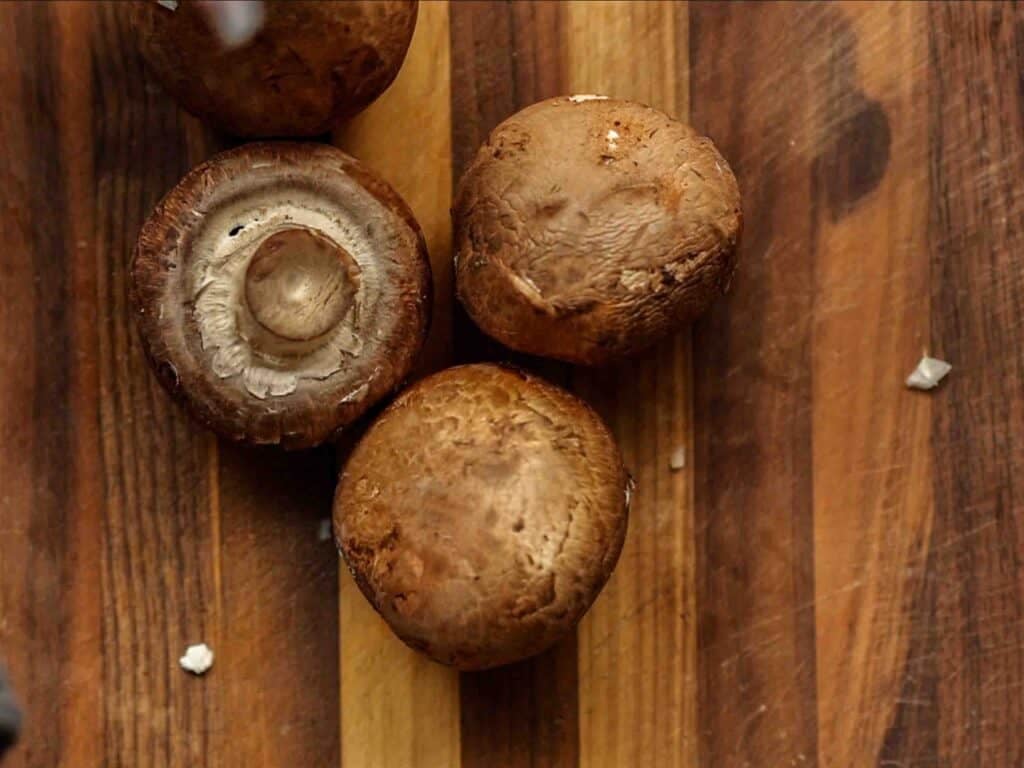 BEST Vegan Bolognese Recipe - Meaty and Delicious. 10 Four whole brown mushrooms lie on a striped wooden cutting board, with one mushroom showing its stem and gills.