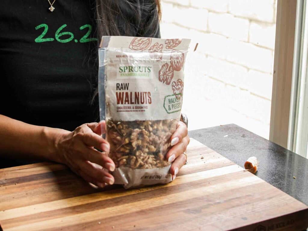 BEST Vegan Bolognese Recipe - Meaty and Delicious. 11 A person wearing a black shirt holds a bag of Sprouts Farmers Market raw walnuts on a wooden cutting board, with some walnut pieces scattered nearby.