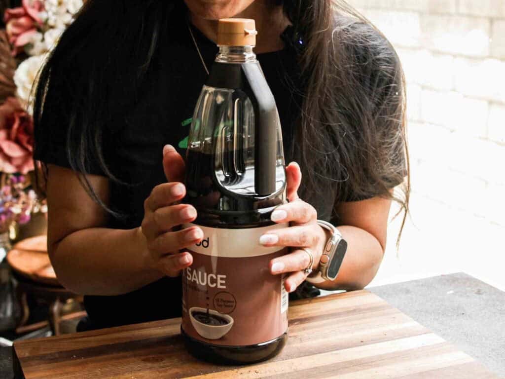 BEST Vegan Bolognese Recipe - Meaty and Delicious. 8 A person with long hair holds a large bottle of soy sauce on a wooden surface. Only their hands and torso are visible. Light comes in from a window in the background.